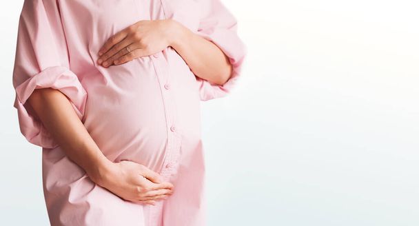 Closeup of beautiful pregnant woman in pink shirt dress holds hands on her belly in white background. Loving mother waiting for baby birth. Pregnancy, maternity, preparation and expectation concept. - Foto, afbeelding