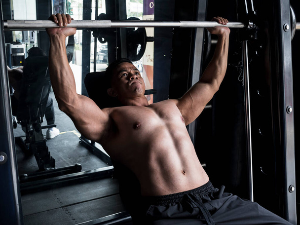 筋肉質なアジア人男性がジムやフィットネスセンターでスミスマシンの傾斜ベンチプレスを実行します。ワークアウトと胸の日のコンセプト. - 写真・画像