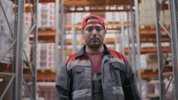 カメラを見て折り畳まれた手で倉庫で追跡パレットの間で全体的な均一で赤いキャップ立っている成人中近東の建設労働者の中スローモーションの肖像画 - 映像、動画
