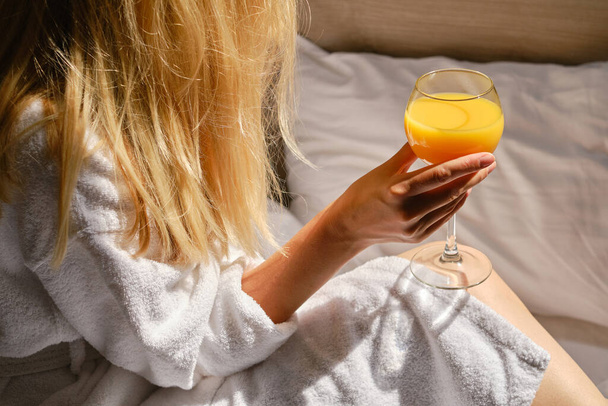 Girl with a glass of orange juice in bed. Vitamin drink before breakfast for happy woman. Orange fresh. A cozy morning on a bed with white linens. Relaxation in the bedroom. Dreams and enjoy in home - Foto, Bild