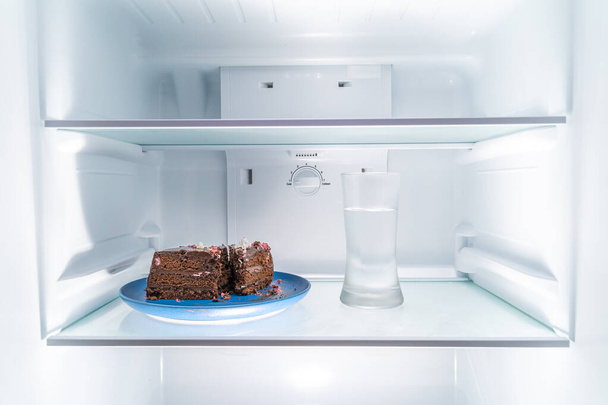 Chocolate cake on a blue plate and a glass of water placed in a clean refridgerator, diet or food concept - Фото, изображение