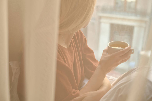 The girl drinks tea in the morning near the window. A cup of hot tea in hands. Awakening from sleep. The happy woman is wrapped in a blanket. Dreams and think about life. Enjoy cozy in home - Foto, afbeelding