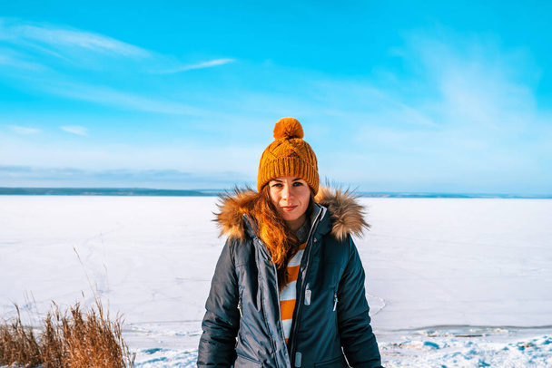 Una donna in giacca invernale e un cappello giallo lavorato a maglia si staglia sullo sfondo di un fiume ghiacciato con un paesaggio invernale - Foto, immagini