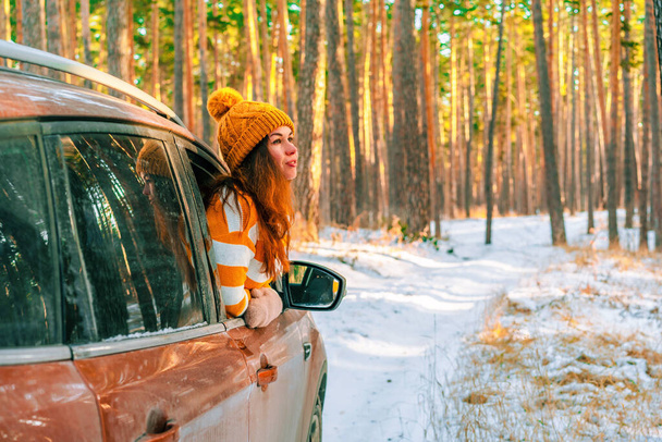 Uśmiechnięta kobieta w swetrze i kapeluszu wyglądająca przez okno samochodu na tle śnieżnego krajobrazu w zimowym lesie sosnowym, podróż - Zdjęcie, obraz