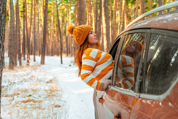 冬の松林の雪景色、ロードトリップを背景に、セーターや帽子の笑顔の女性は車の窓から外を見る - 写真・画像