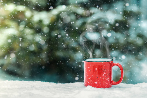 A red cup with a hot winter drink (mulled wine, cocoa, coffee, tea) against the background of a winter forest and snow. Winter mood and comfort. - Foto, Imagem