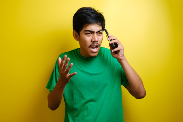 Closeup portrait angry young asian man, guy mad student, pissed off employee shouting while on phone isolated yellow wall background. Negative human emotion face expression feeling attitude - Photo, Image