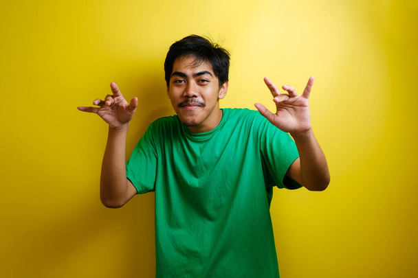 Homem asiático em camiseta verde sorrindo e dançando feliz, alegre expressando celebrando boa notícia vitória ganhando gesto de sucesso contra fundo amarelo - Foto, Imagem