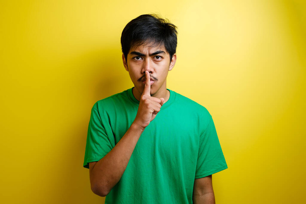 Yeşil tişörtlü Asyalı adam izole edilmiş sarı arka planda sessizlik hareketi yapıyor. - Fotoğraf, Görsel