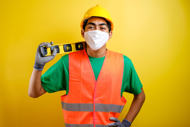 Maske ve güvenlik yeleği takan Asyalı inşaat işçisi kendine güvenini göstermek için omzunda şelale tutuyor. Sarı arkaplana karşı çalışma kavramına hazır - Fotoğraf, Görsel