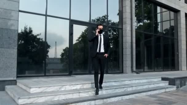 Een man in een pak, die aan de telefoon praat, loopt van een gebouw met glazen ramen naar zijn individuele milieuvriendelijke vervoer. - Video