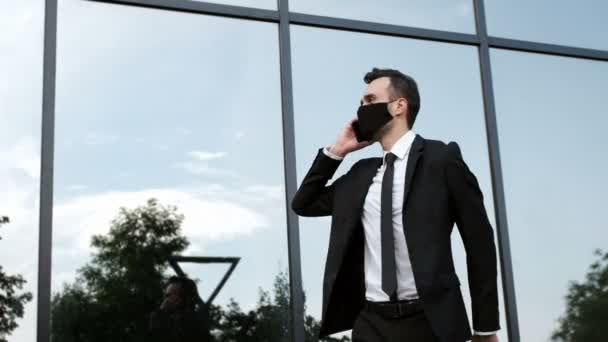 Een man in een zwart pak en een zwart masker communiceert zenuwachtig op een smartphone, kijkt naar zijn horloge, stampt ter plekke, maakt zijn das los. - Video