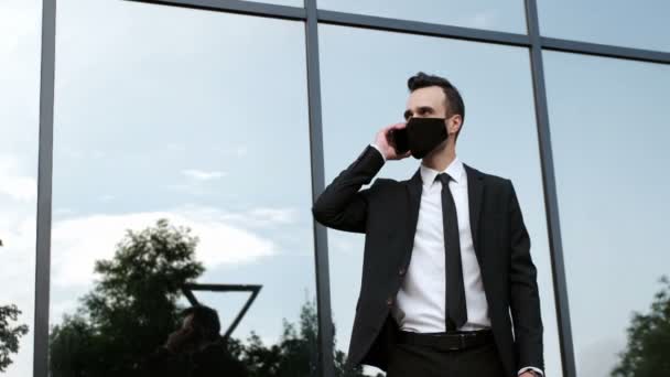 Een zakenman met een zwart masker beantwoordt een telefoongesprek scherp, kijkt rond en kijkt hoe laat het is. Een zakenman in quarantaine voert een zakelijk gesprek. - Video