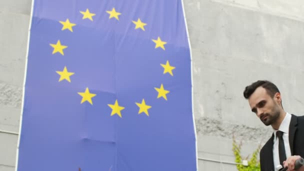 Un homme en tenue de travail entre dans le cadre avec un scooter sur le fond du drapeau de l'Union européenne, regarde le drapeau et sourit optimiste.  - Séquence, vidéo