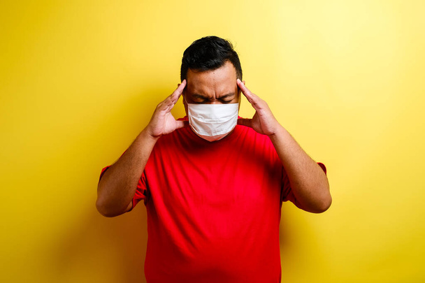Portrait of young ill man in medical protective mask on face suffering from headache and weakness, isolated on yellow background - Valokuva, kuva