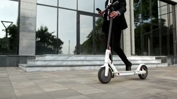 A man in a business suit looks at his smartphone and rolls an electric scooter next to him against the background of large glass windows. - Footage, Video