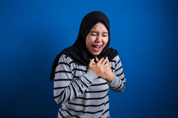 Asian muslim college student girl feeling pain in her chest, gesture holding left chest. Blue background - Foto, Imagem