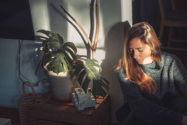 Beautiful young woman next to a trendy monstera plant with big leaves indoor with morning sun rays. Autumn concept. - Zdjęcie, obraz