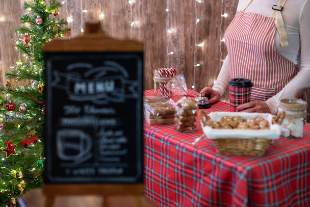 Christmas pastry shop . Woman seller, waitress selling gingerbread, marshmallows, cookies, sweets and hot chocolate in a small cozy cafe. Homemade bakery menu in the foreground - Foto, imagen