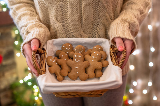 Woman in pullover holding straw basket with gingerbread man on the christmas tree background with beautiful light bokeh. Copy space. - Foto, Imagem