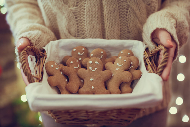 Woman in pullover holding straw basket with gingerbread man on the christmas tree background with beautiful light bokeh. Copy space. - Φωτογραφία, εικόνα