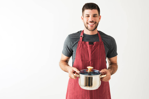 Portret van een lachende jongeman met een schort en een kookpot met soep voor een witte achtergrond - Foto, afbeelding