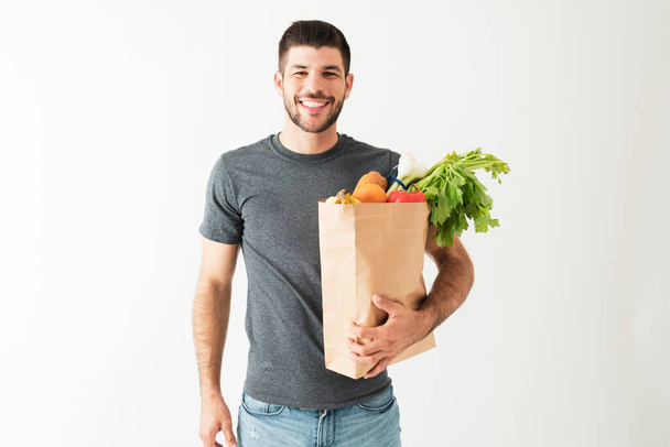 Un giovane ispanico sorridente sta trasportando una borsa della spesa con frutta e verdura fresca, pronta per iniziare una dieta sana. - Foto, immagini