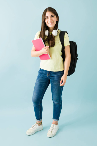 Visão completa de uma jovem estudante sorridente com fones de ouvido, carregando um caderno e uma mochila. Isolado contra um fundo azul - Foto, Imagem