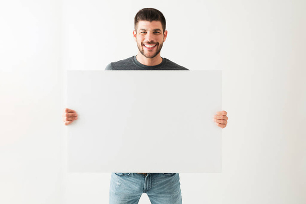 Ritratto di un giovane ispanico che sorride e sembra felice mentre tiene un cartello bianco bianco su uno sfondo bianco - Foto, immagini
