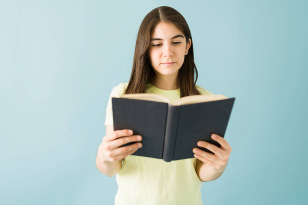 Young female student holding a book and reading with an interested look in front of a blue background - Foto, Imagem