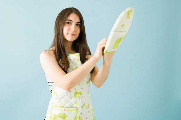 Portrait of an attractive caucasian woman wearing an apron and putting on kitchen gloves in front of a blue background - Photo, Image