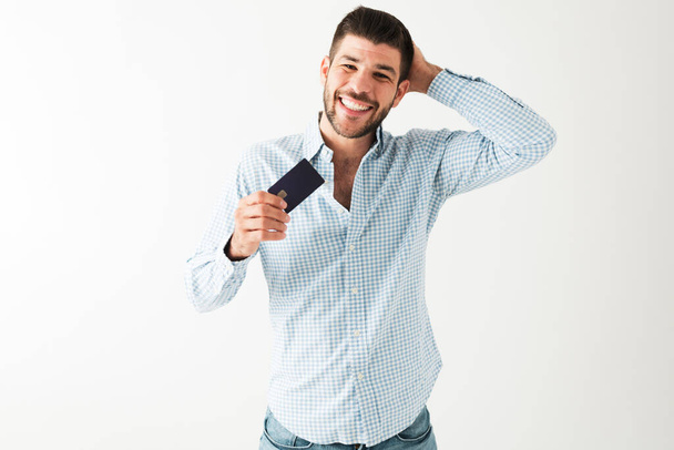 Handsome young guy in his 20s is smiling and looking very happpy, holding a credit card with a hand behind his head - Photo, Image