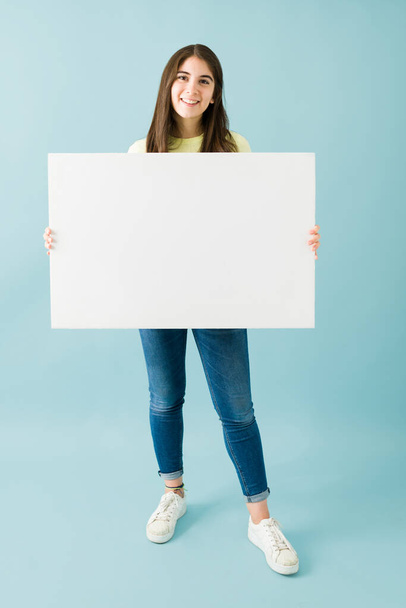 Caucasian young woman smiling and looking happy while holding a white blank sign against a blue background - Zdjęcie, obraz