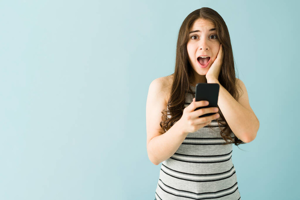 Biała młoda kobieta trzyma smartfona z zaskoczonym i zszokowanym wyrazem twarzy na tle niebieskiego tła - Zdjęcie, obraz