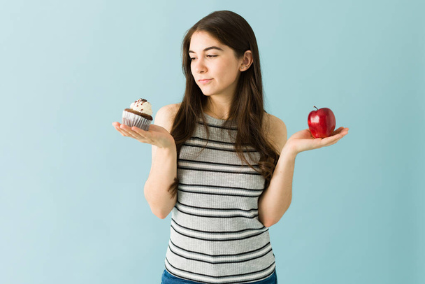 Όμορφη νεαρή γυναίκα που κρατάει ένα μήλο και ένα κεκάκι σοκολάτας σε κάθε χέρι και αποφασίζει τι θα φάει. Γυναίκα σε μια υγιεινή διατροφή - Φωτογραφία, εικόνα