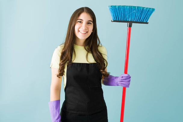 Attractive young woman with an apron and gloves. Woman holding a broom and about to do housework - Photo, Image