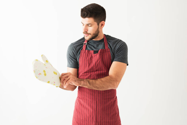 Retrato de un hombre atractivo usando un delantal y poniéndose guantes de cocina frente a un fondo blanco - Foto, imagen