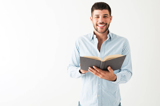 Der hispanische junge Mann trägt ein Hemd, lächelt in die Kamera und hält ein aufgeschlagenes Buch vor weißem Hintergrund - Foto, Bild