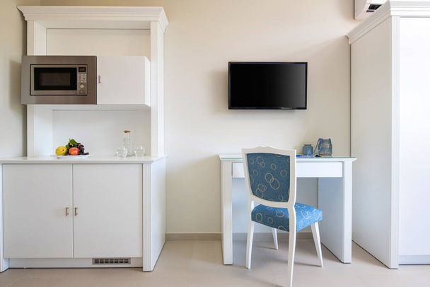 テレビ、テーブル、古典的な青の椅子、小さな部屋のモダンなインテリアで組み込みの家電付きの木製キッチン食器棚付きの白い壁の正面ビュー - 写真・画像