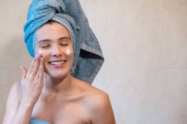 Młoda kobieta uśmiechnięta w łazience podczas stosowania kremu nawilżającego na twarz z ręcznikiem zwiniętym na głowie. Pojęcie pielęgnacji skóry podczas porannej rutyny. - Zdjęcie, obraz