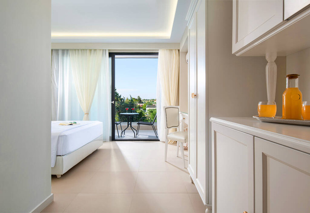 Ontspanning concept van klassieke stijl interieur resort hotel appartement met welkom sinaasappelsap drankje. Witte slaapkamer grote open raam - glazen deur naar de zomer prachtig landschap - Foto, afbeelding