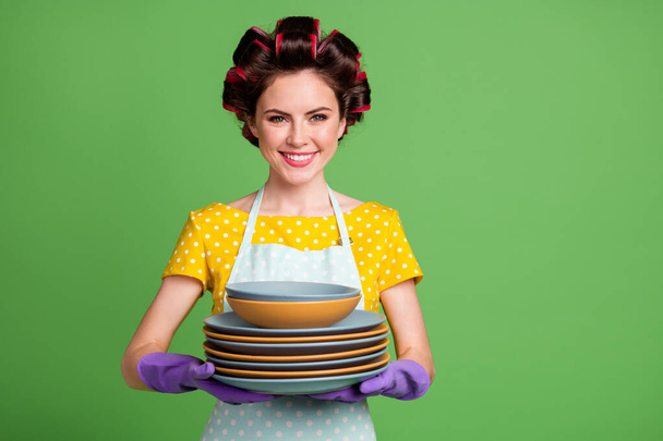 Photo of positive maid girl wash dishes after family party hold pile stack wear yellow dotted dress hair rollers isolated over green color background - Foto, Bild