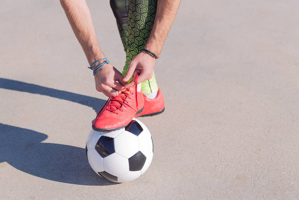 detail of a football player tying his boots with a foot on the ball in a concrete soccer field, concept of healthy lifestyle and urban sport in the city, copy space for text - Photo, image