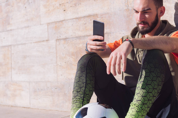 handsome football player rest sitting on the court consulting his smart phone, concept of technology and urban sport lifestyle in the city, copy space for text - Photo, Image