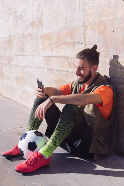 vertical photo of a fashionable soccer player rest sitting on the court consulting his mobile phone, concept of technology and urban sport lifestyle in the city, copy space for text - Foto, afbeelding