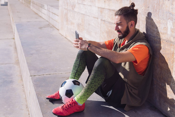 fashionable football player rest sitting on the court consulting his mobile phone, concept of technology and urban sport lifestyle in the city, copy space for text - Fotografie, Obrázek
