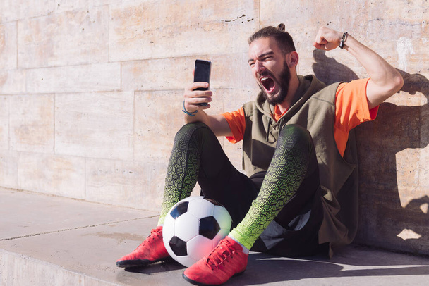 hipster football fan celebrates a victory when he is checking his smart phone, concept of technology and urban sport lifestyle in the city, copy space for text - Zdjęcie, obraz