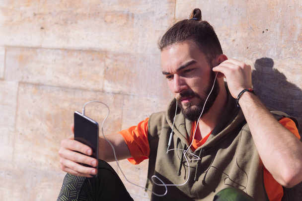 handsome sportsman putting on earphones to listen to music from his smart phone, concept of technology and urban sport lifestyle in the city, copy space for text - Photo, Image