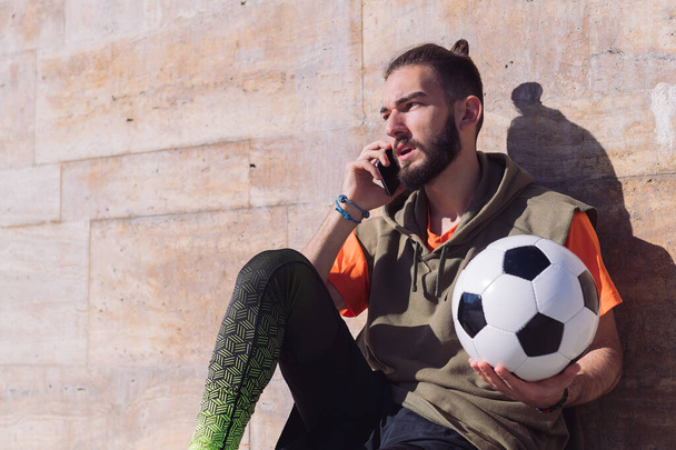 handsome sportsman talking on the phone while resting with his football ball, concept of technology and urban sport lifestyle in the city, copy space for text - Photo, Image