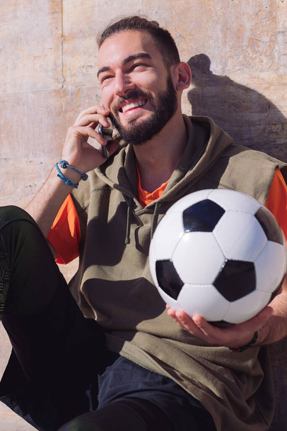handsome sportsman laughing and talking by phone while resting with his football ball, concept of technology and urban sport lifestyle in the city - Fotografie, Obrázek
